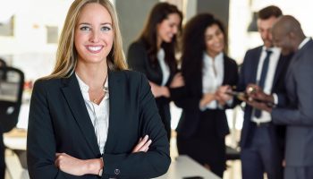 Businesswoman leader looking at camera in modern office with multi-ethnic businesspeople working at the background. Teamwork concept. Blonde caucasian woman